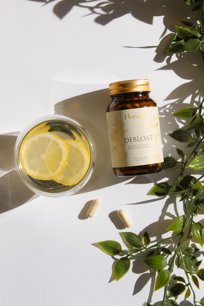 A bottle of Floraelle Debloat with its cap resting beside it, next to a glass of water with lemon slices and trailing green leaves, in soft daylight.